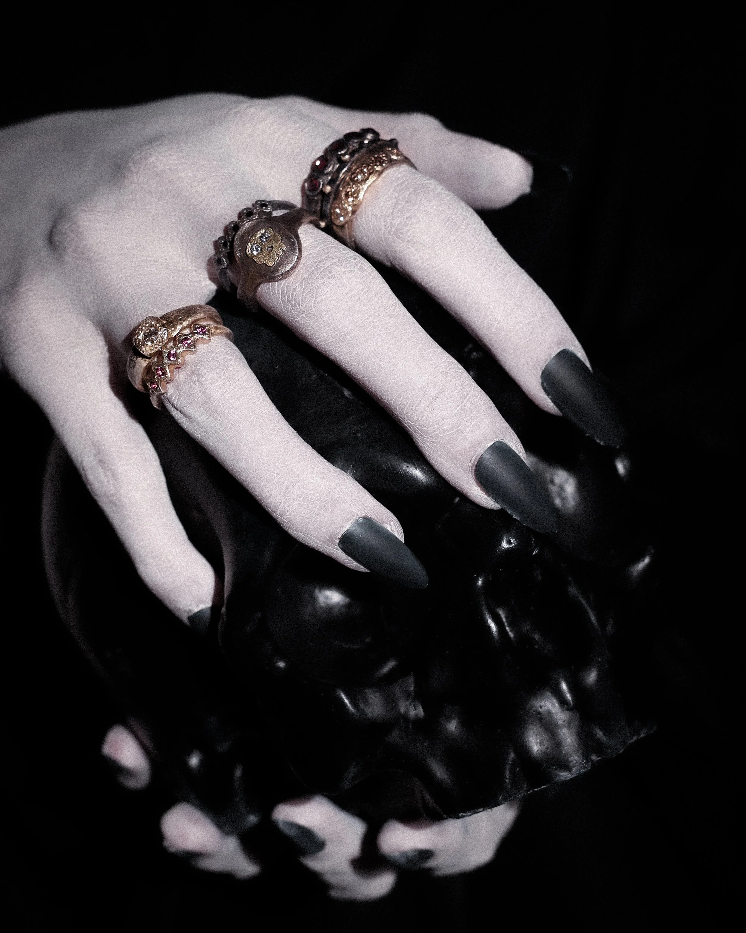 Close-up of a hand with black painted nails wearing multiple silver rings with skull and gemstone designs, against a black background.