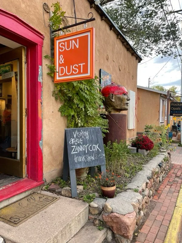 Exterior scene of a quaint shop featuring a bright orange sign that reads 'SUN & DUST', a chalkboard sign advertising a visiting artist Zanny Cox, a bust sculpture with a red headband, and various potted plants with flowers along a brick sidewalk.