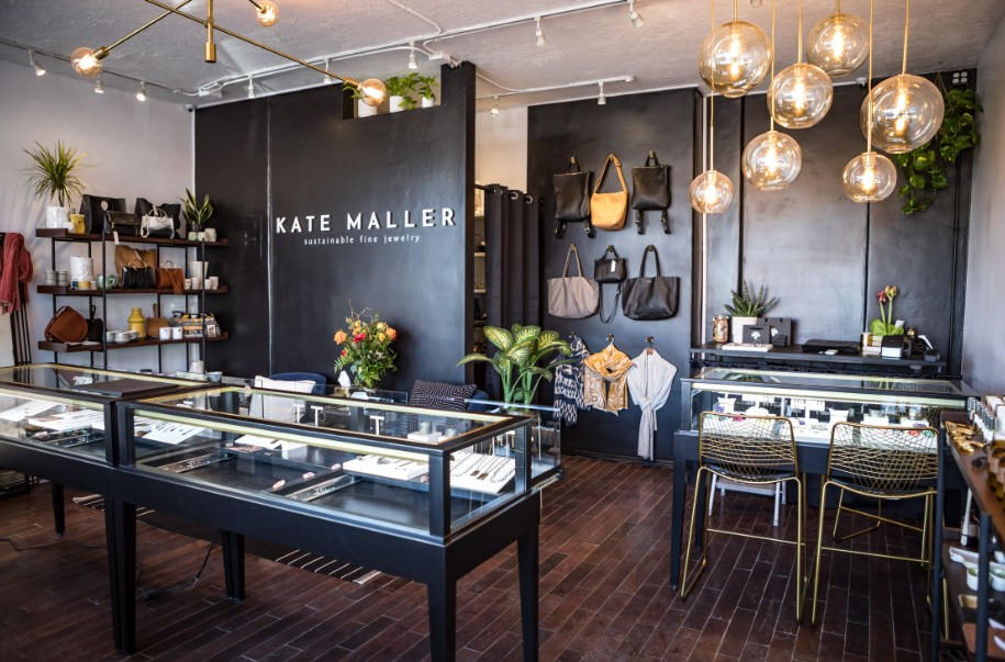 Interior of a jewelry store named Kate Maller with display cases, wall shelves, handbags, and jewelry on tables, with modern lighting and minimalist decor.