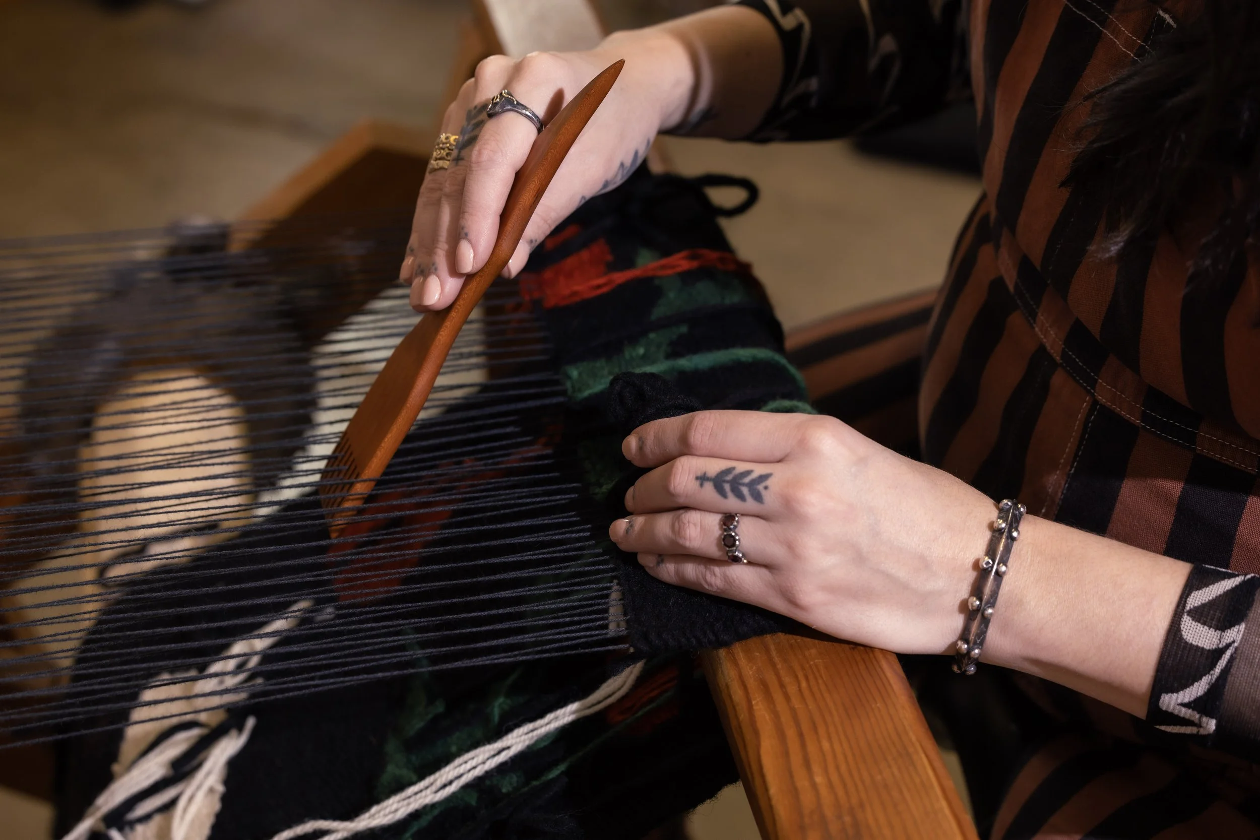 Person weaving on a loom, with hands adjusting the threads, wearing rings and bracelets, with tattoos on fingers, and a striped shirt.