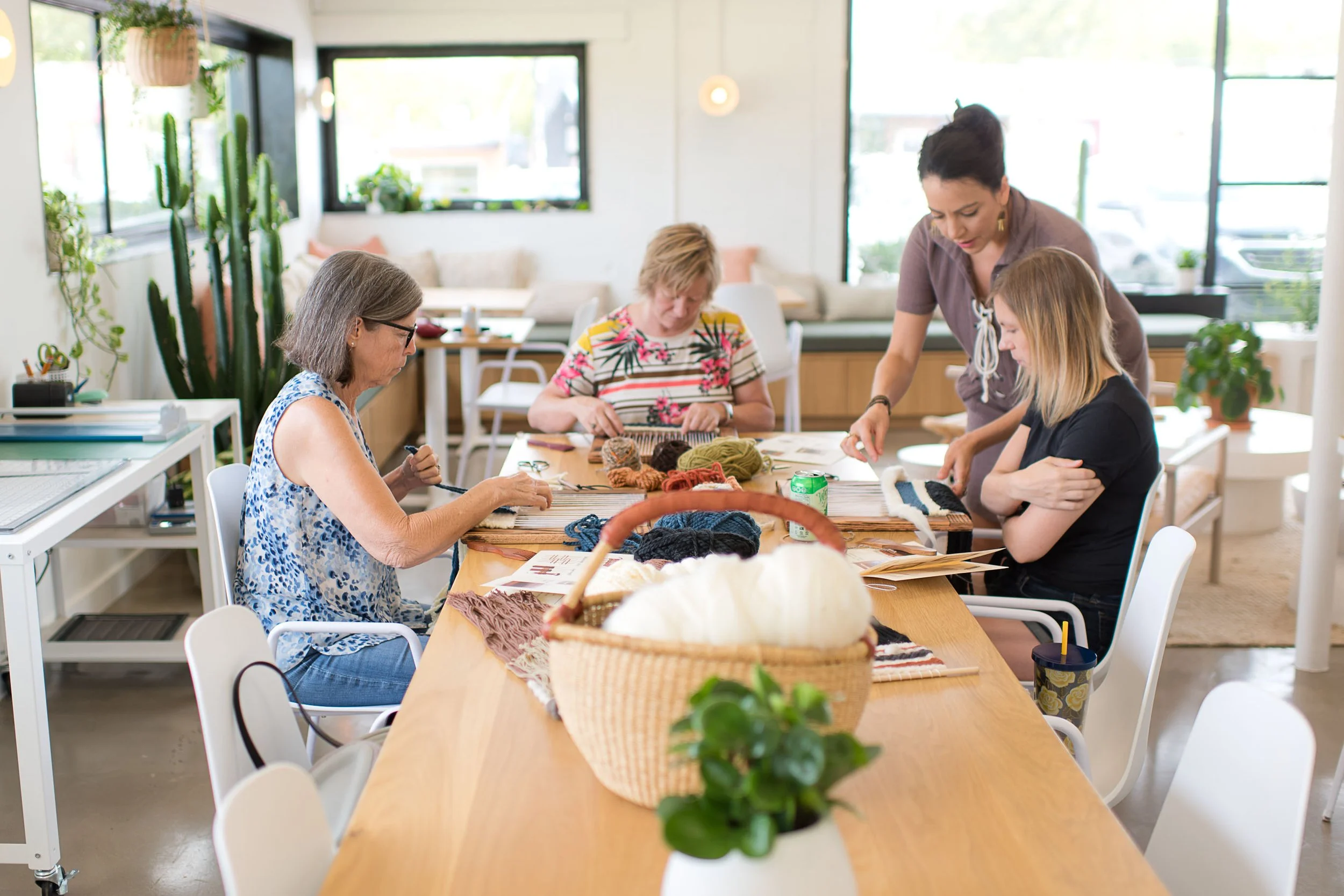 Five women craft with yarn and knitting needles at a bright, cozy table in a room decorated with houseplants and large windows.
