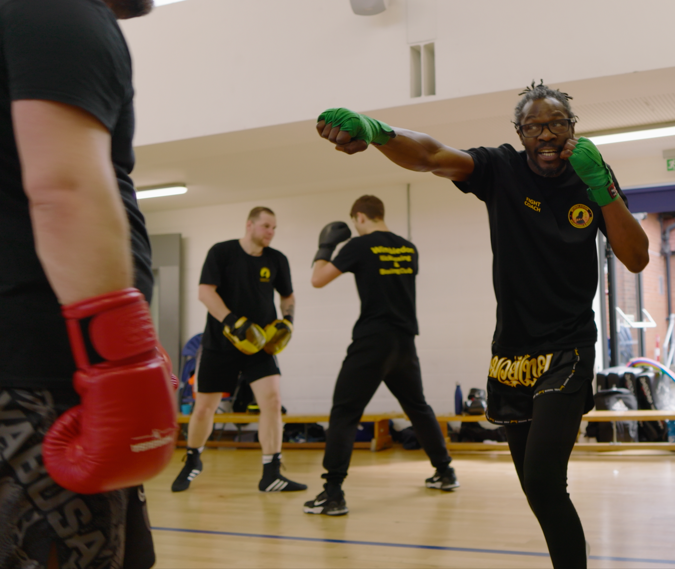 Group boxing training session with a focus on a male coach demonstrating a punching technique. Participants are wearing black shirts, and one person has red boxing gloves.