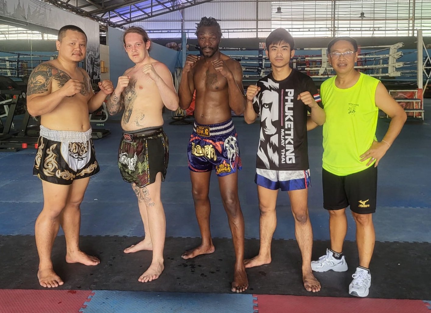 Five men posing in a martial arts gym, wearing Muay Thai shorts and some with tattoos, in front of boxing rings.