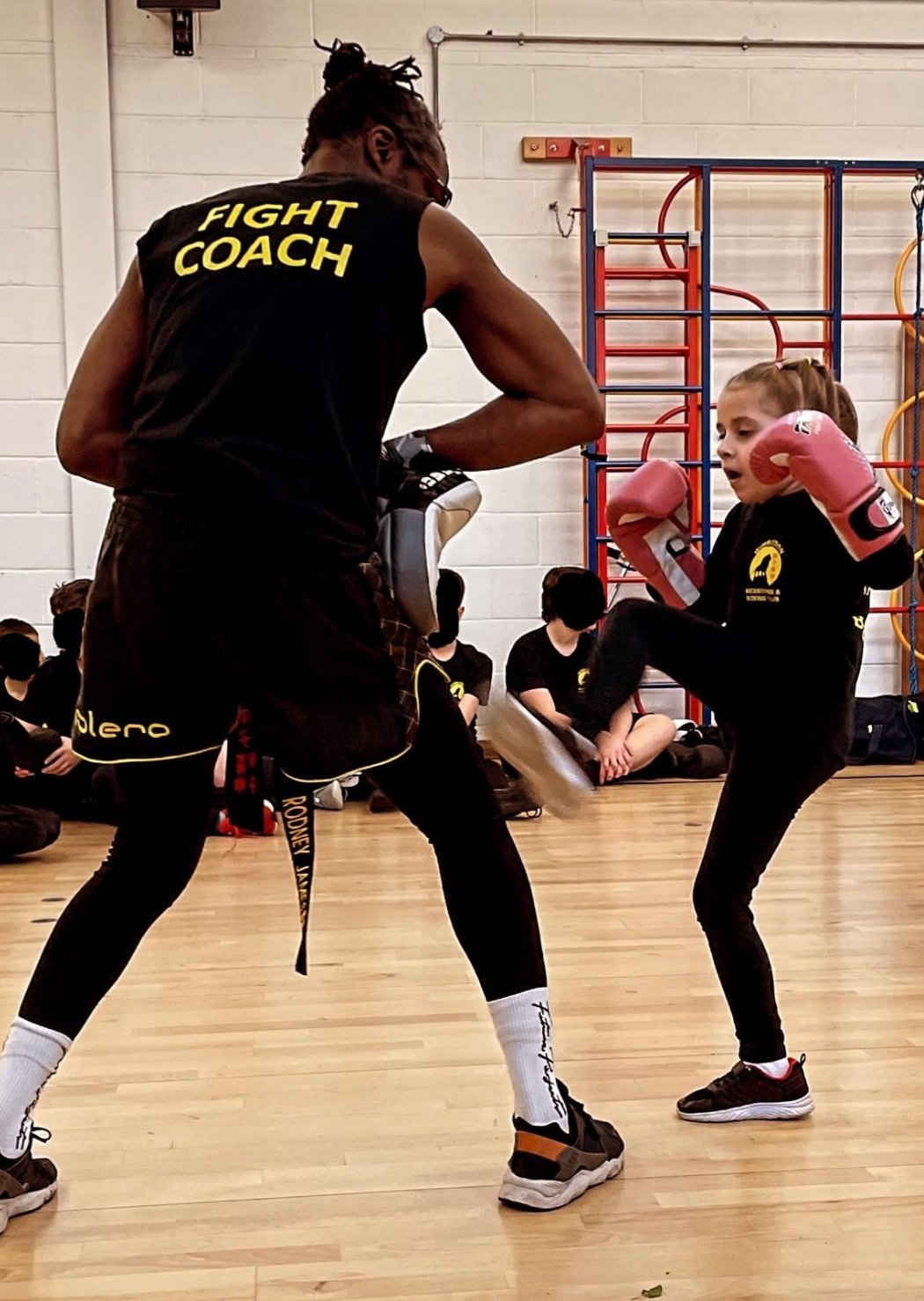 A young girl practices kickboxing with a coach in a gymnasium. The coach wears a "Fight Coach" tank top and holds pads for training. The girl wears gloves and is performing a kick. Several individuals are seated in the background watching.