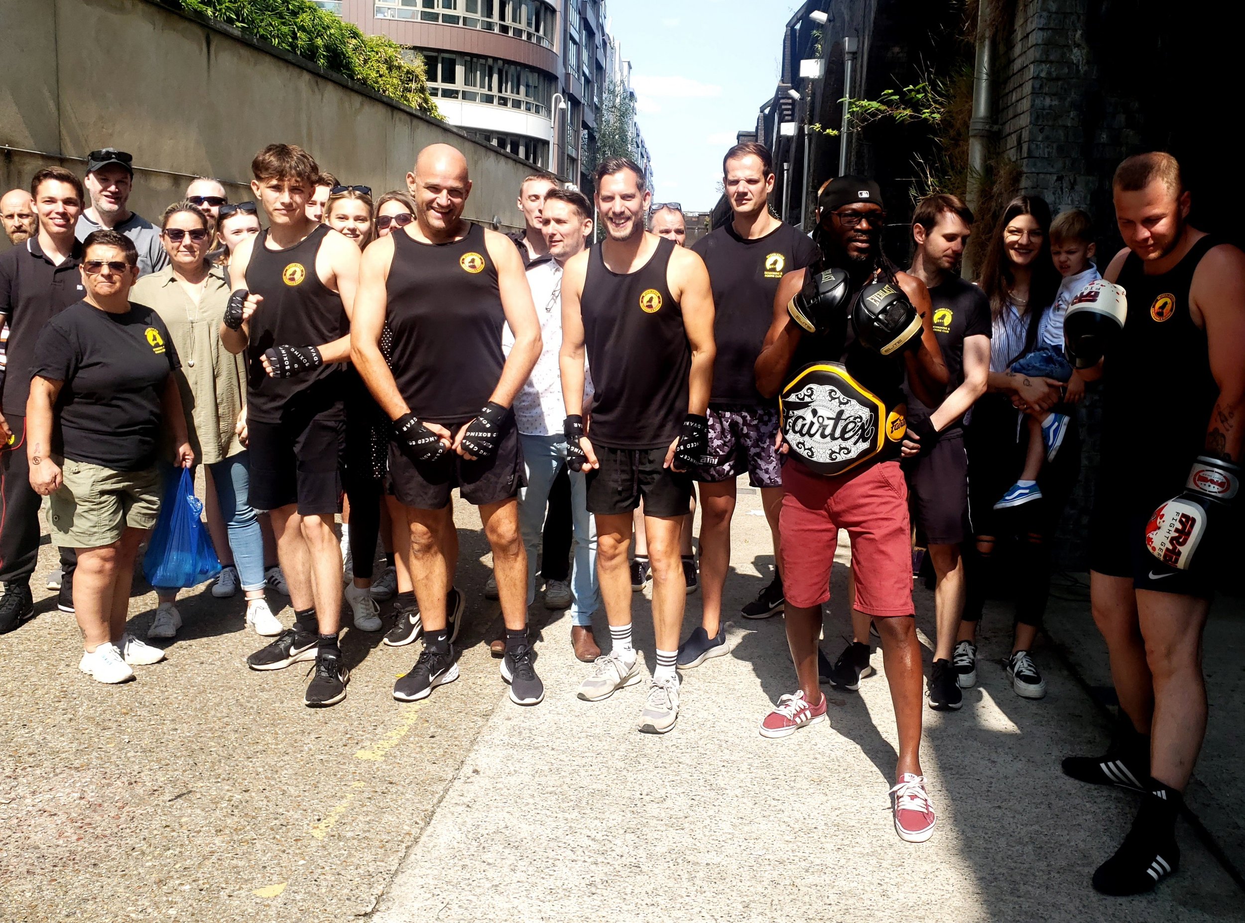 Group of people standing on a street, some in athletic gear, others in casual clothing, one person holding a championship belt.