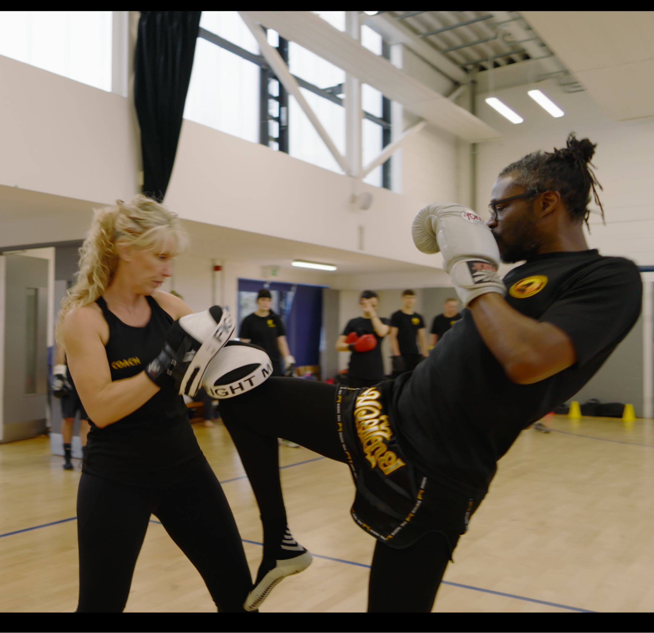 A sparring session in a gym with a man and woman practicing martial arts. The woman is wearing a tank top labeled 'Coach' and has training pads, while the man is performing a high kick, both wearing protective gear. A group of people watches in the b