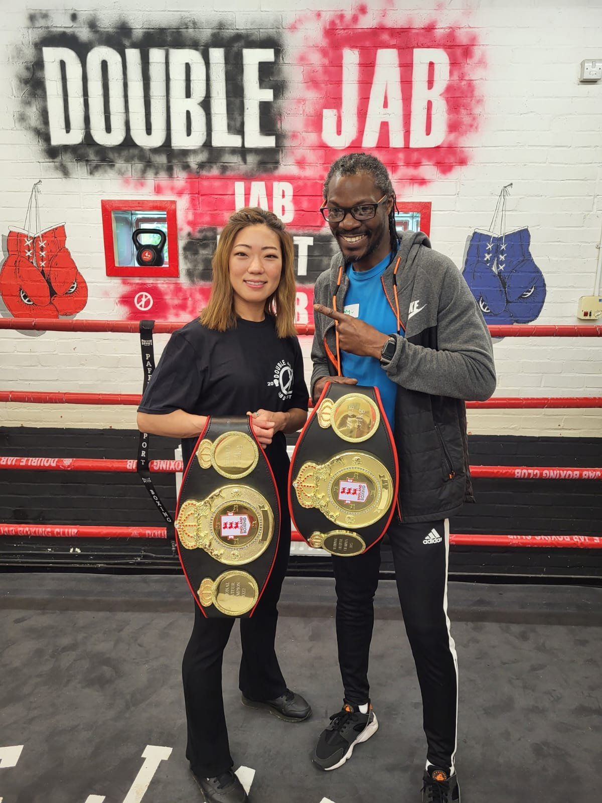 Two people posing with championship belts at a gym, with a wall sign reading 'Double Jab' and boxing gloves artwork in the background.