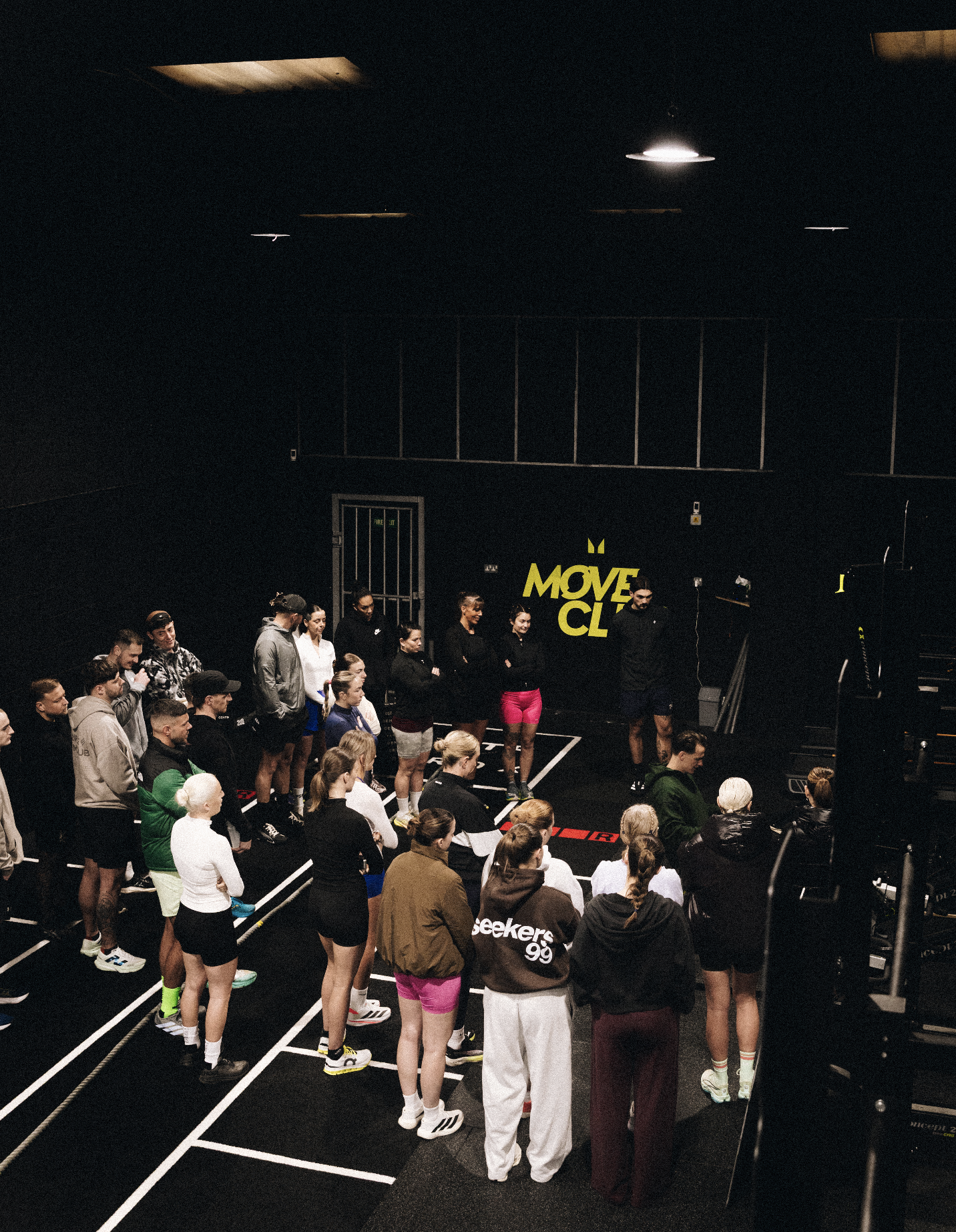 People standing in a lined formation on indoor gym or studio floor with black walls, bright ceiling lights, and a wall-mounted yellow sign reading "MOVE CL".