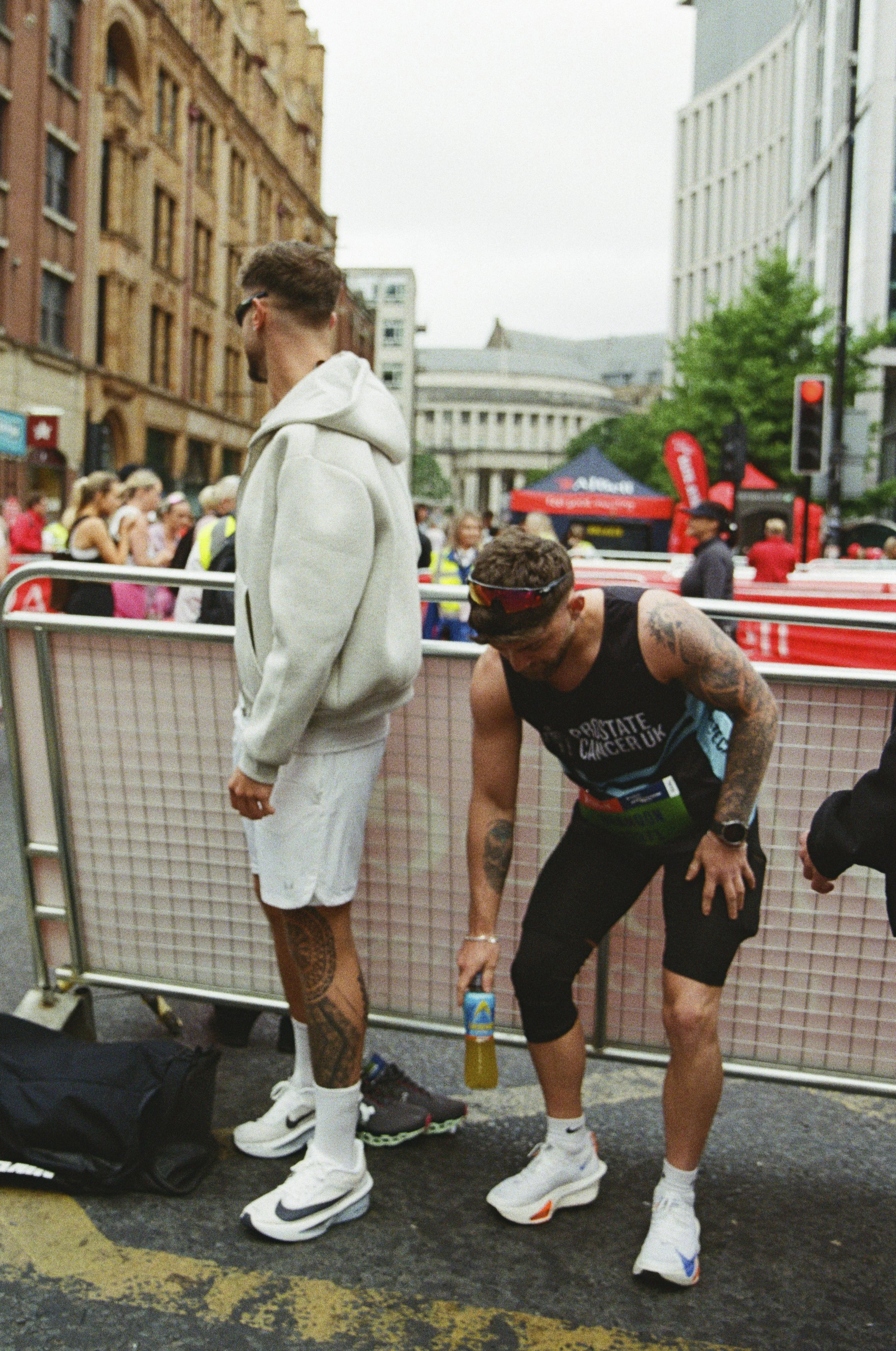 Two male runners are preparing for a race, standing behind a barrier with a crowd and city buildings in the background. One runner is standing while the other is bent over, adjusting something on his leg.