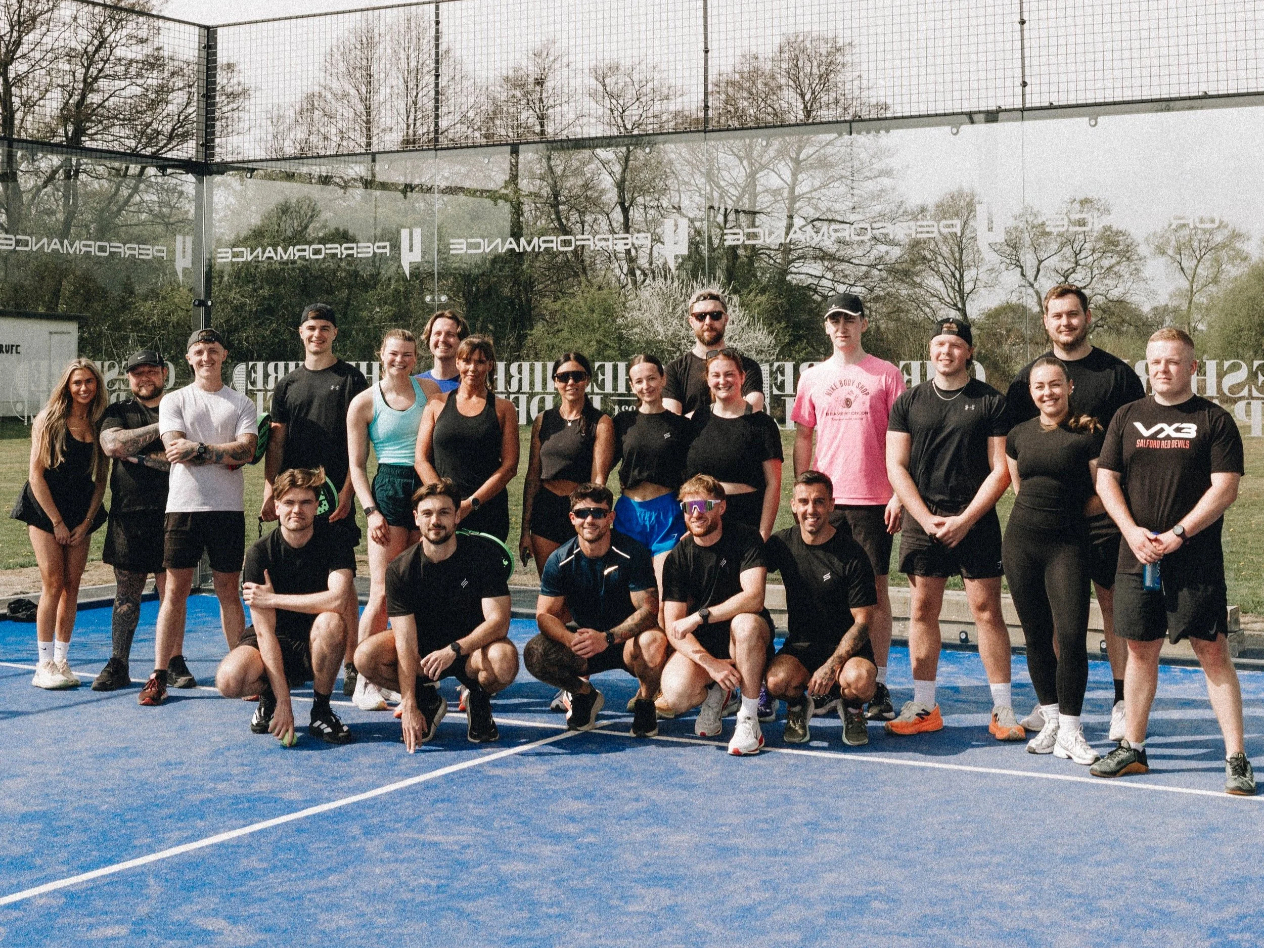 A group of people, mostly in athletic clothing, posing on a blue tennis or sports court outdoors. Trees are visible in the background.