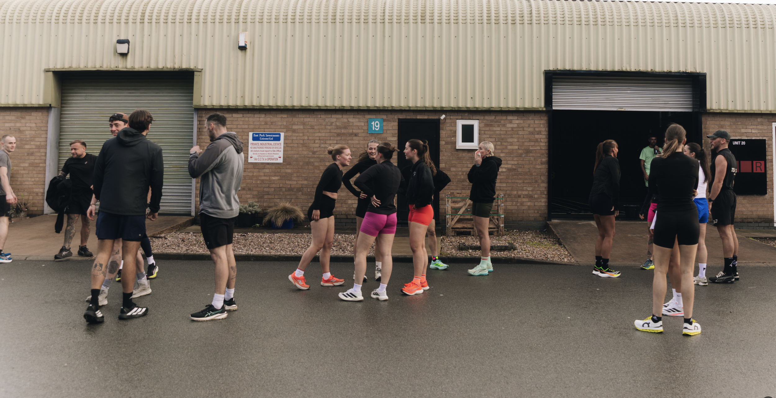 Group of people dressed in athletic wear standing outside a brick industrial building, talking and socializing.