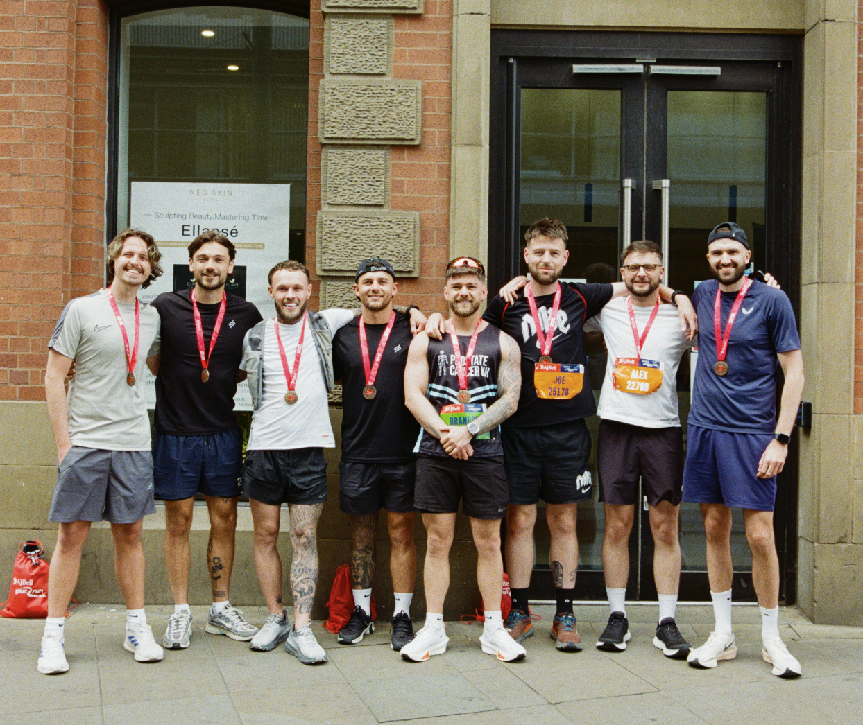Group of eight men standing outside in front of a building, wearing running clothes and medals, smiling, after participating in a race or marathon.