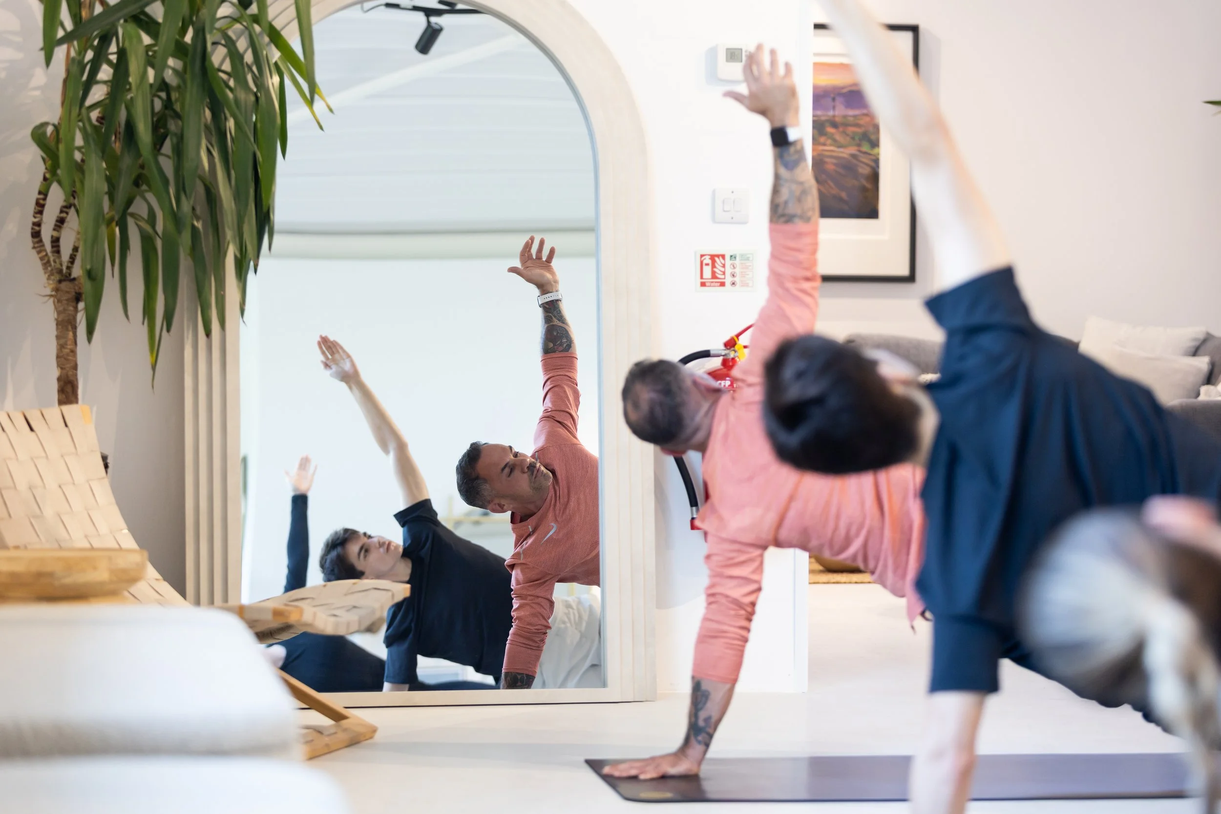 People participating in a yoga or stretching class inside a room with a mirror, fire safety signs, and a fire extinguisher.