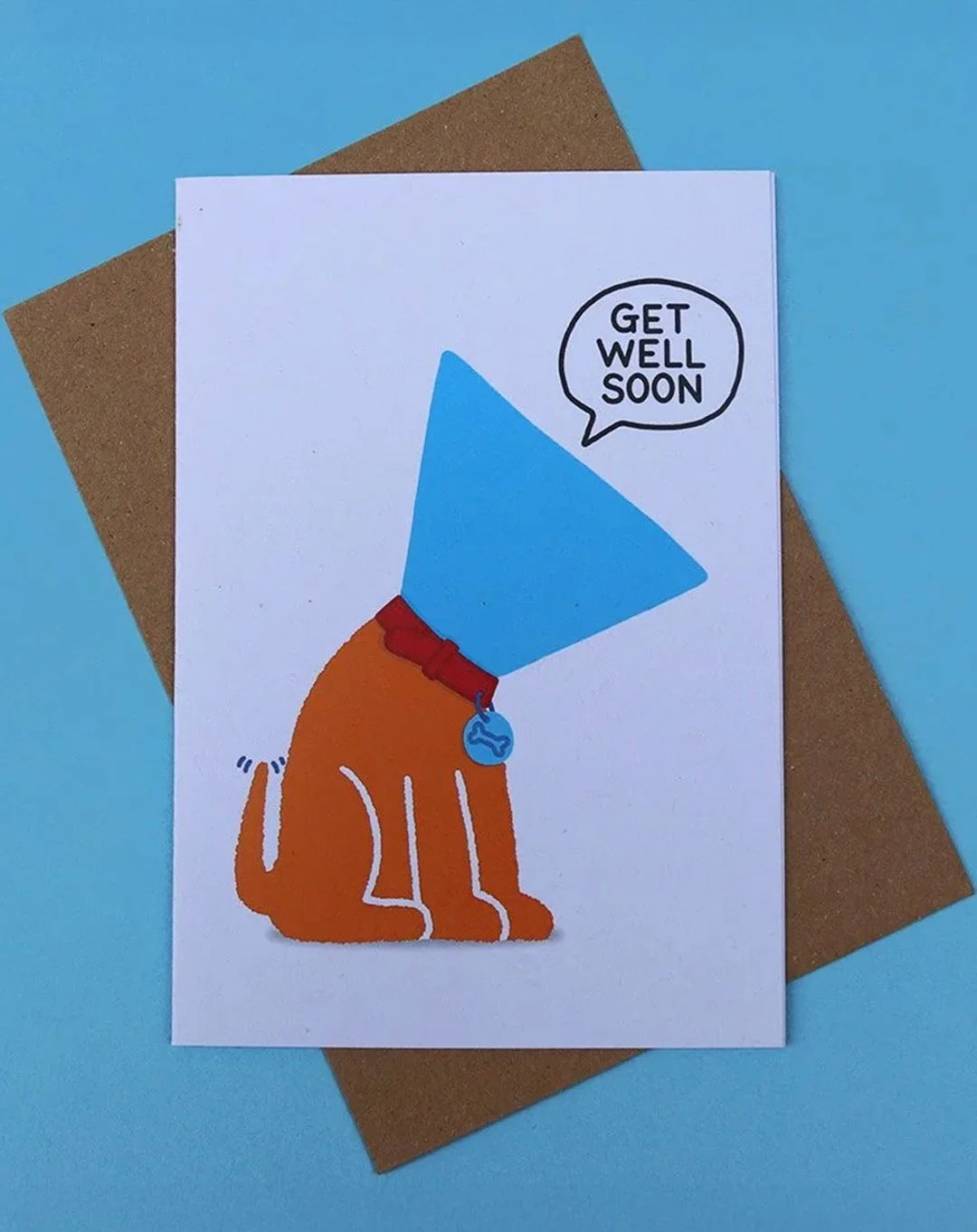 Greeting card with "Get Well Soon" text on an white background, featuring a dog wearing a cone of shame designs, on a blue surface, with a kraft envelope.