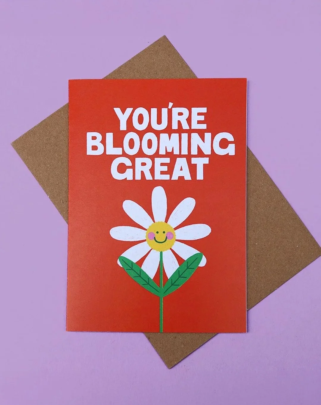 Greeting card with "You're Blooming Great" text on an orange background, featuring a daisy flower character designs, on a lilac surface, with a kraft envelope.