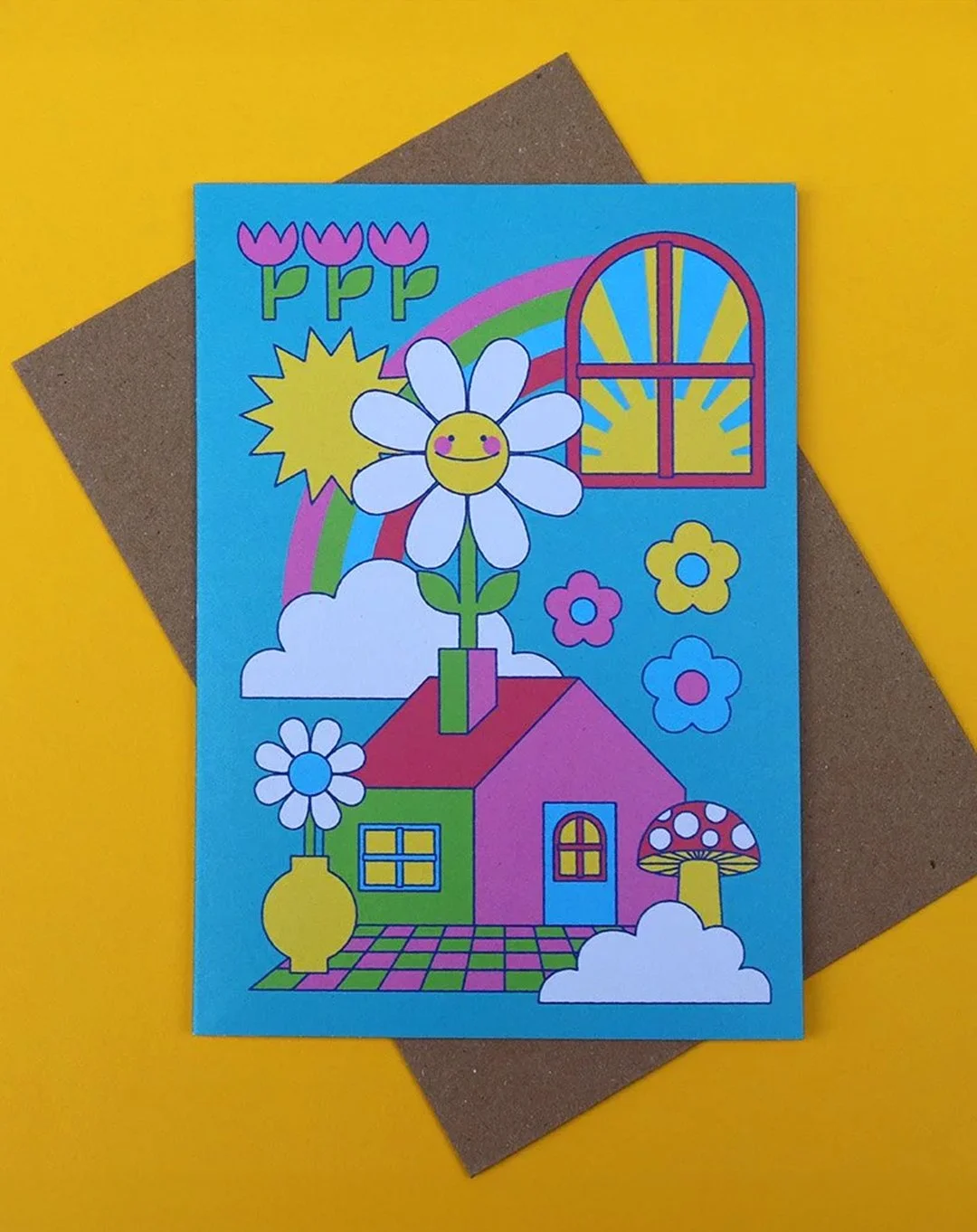Greeting card featuring a graphic style design, with a house blooming a daisy happy flower from the chimney, including mushroom, flowers and vase, as well as a window with a sun. All on a blue background. The card is on a yellow surface, with a kraft