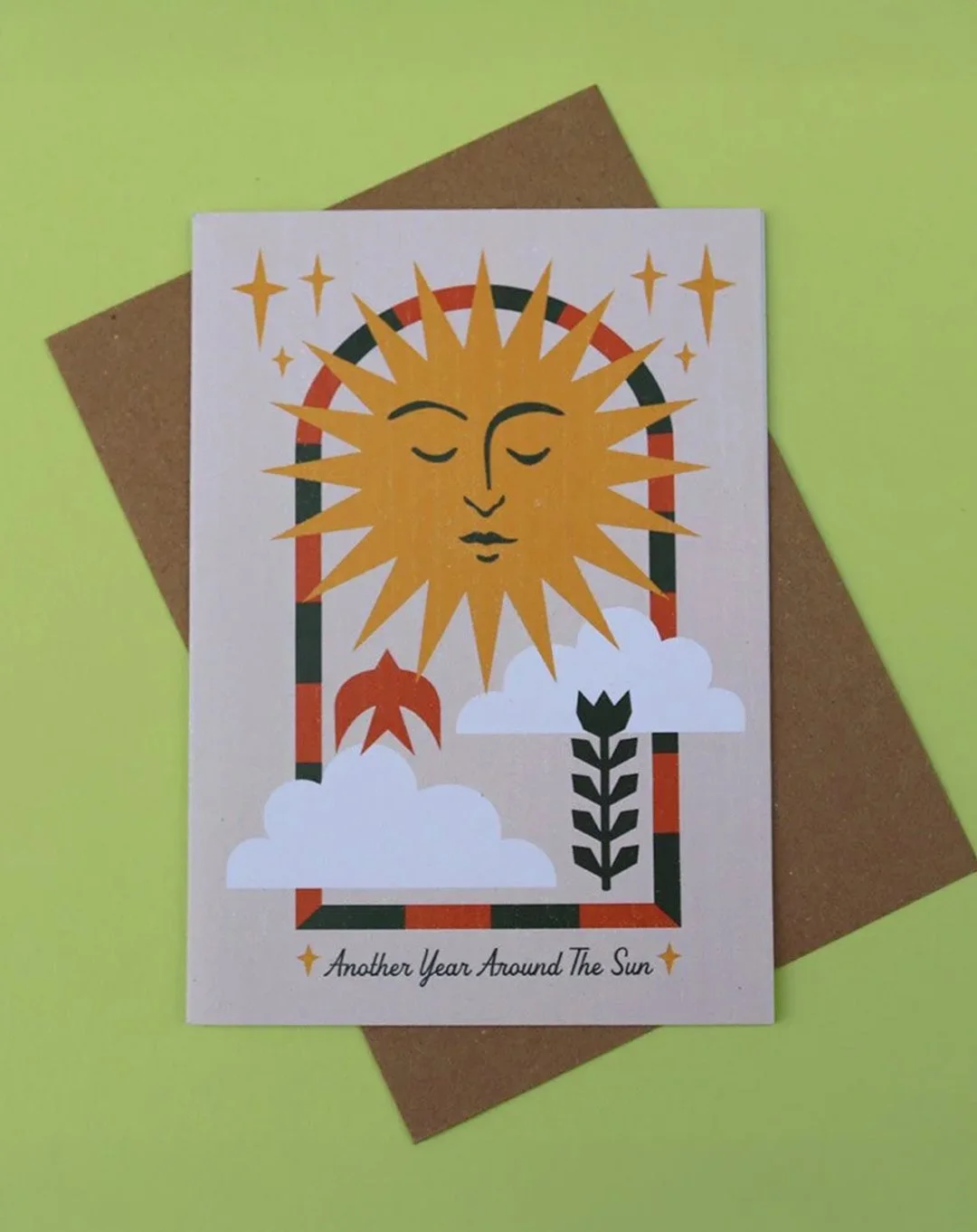 Greeting card with "Another Year Around The Sun" text on an beige background, featuring a sun celestial designs, on a green surface, with a kraft envelope.