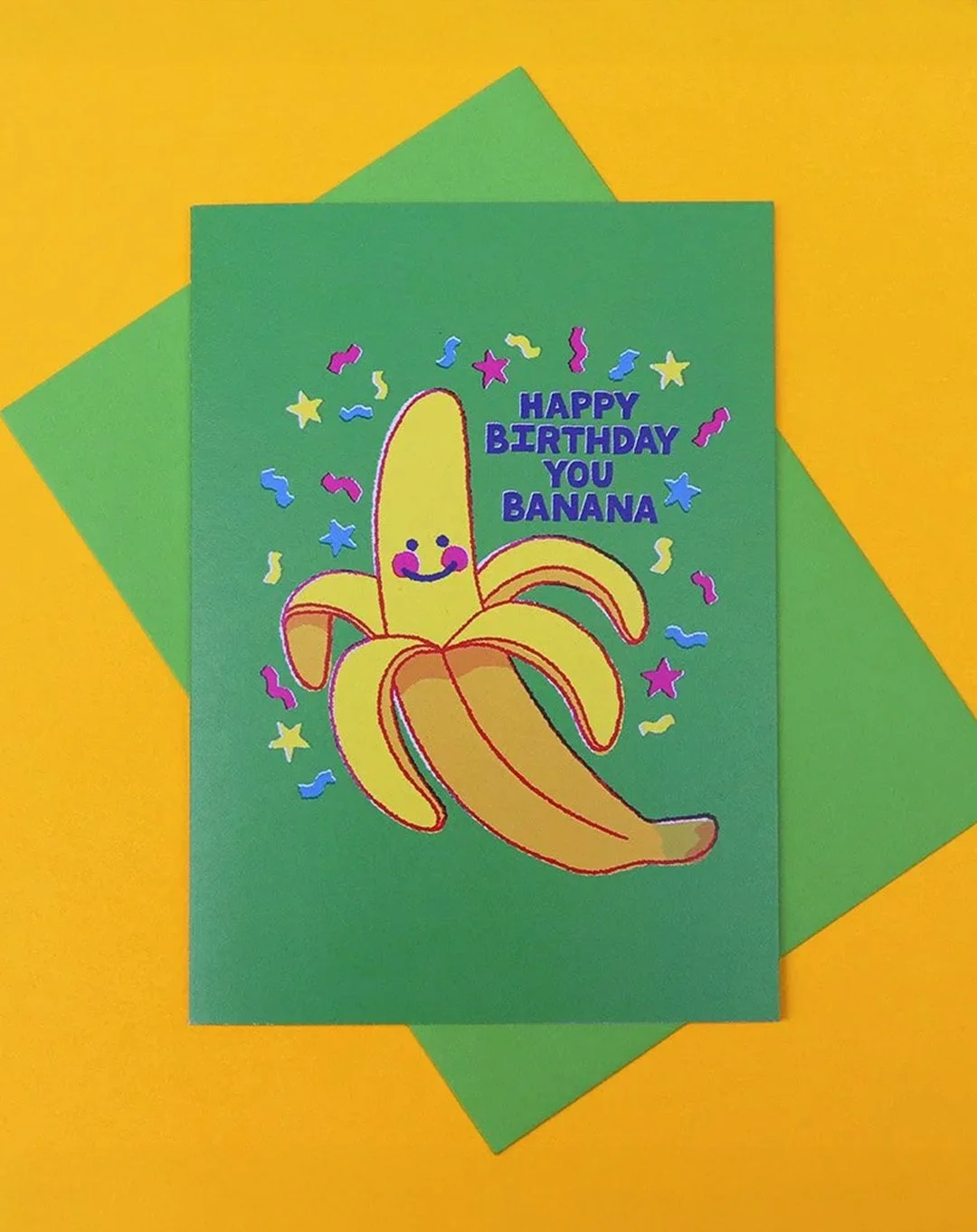 Greeting card with "Happy Birthday You Banana" text on a green background, featuring a happy banana character and confetti designs, on a yellow surface, with a green envelope.