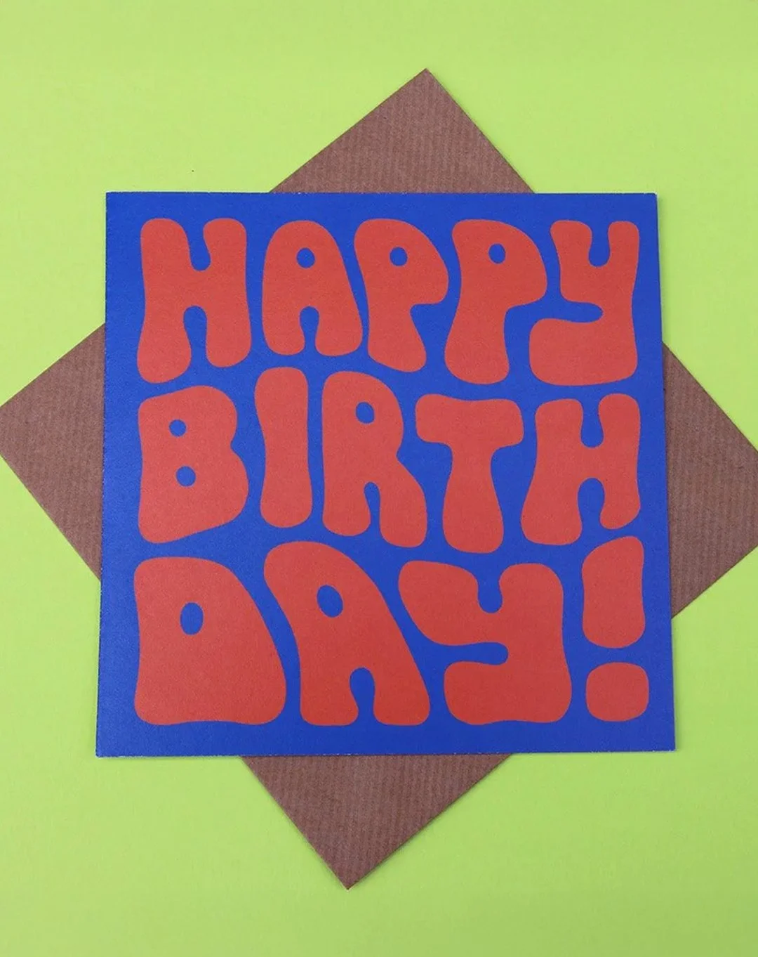Greeting card with orange "Happy Birthday!" bubble text on a blue background, on a green surface, with a kraft envelope.