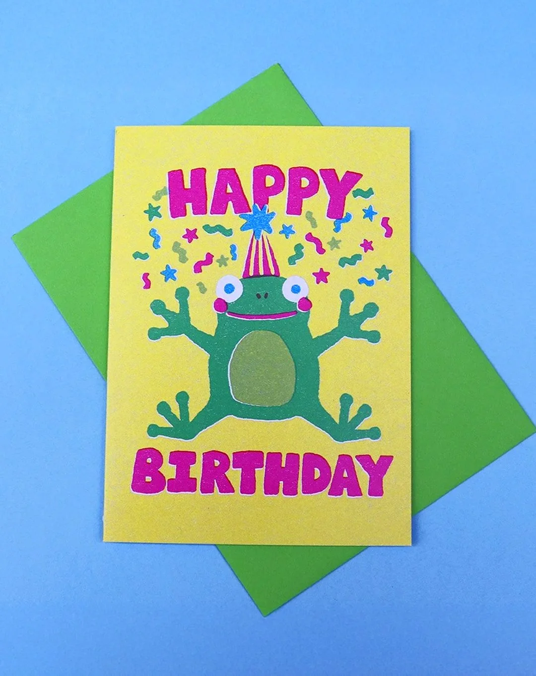 Greeting card with "Happy Birthday" text on yellow background, featuring a leaping frog and confetti designs, on a blue surface, with a green envelope.