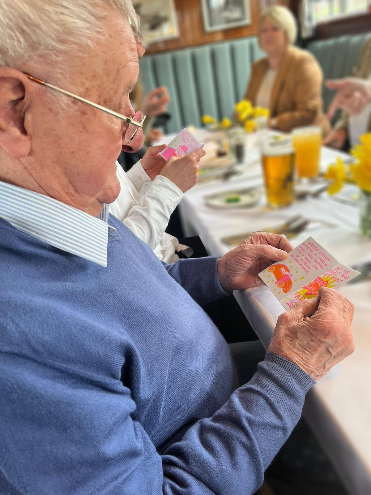 Elderly man (Grandad) reading a 19333 zine at a dining table, with flowers and drinks in the background.