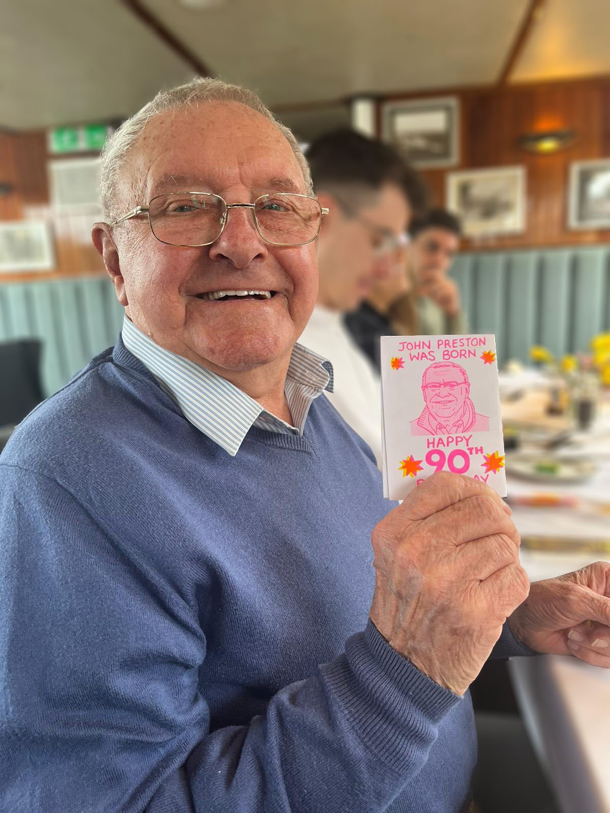 Smiling elderly man (Grandad) holding a 90th birthday zine at a dining table indoors.