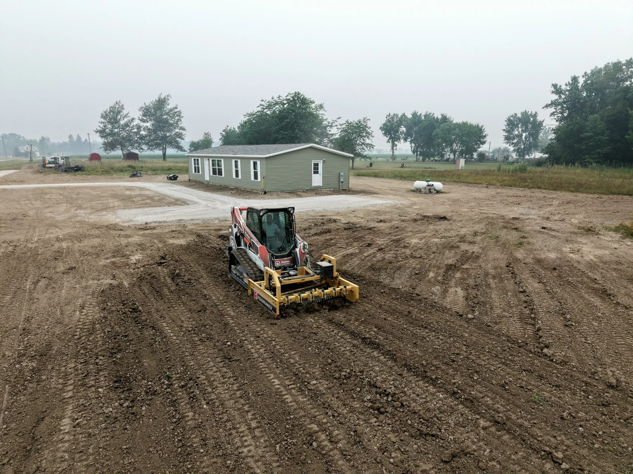 Construction site with dirt ground, a small gray building, and a yellow skid-steer loader in foreground. There are trees, utility poles, and some storage sheds in the background, with a cloudy sky.