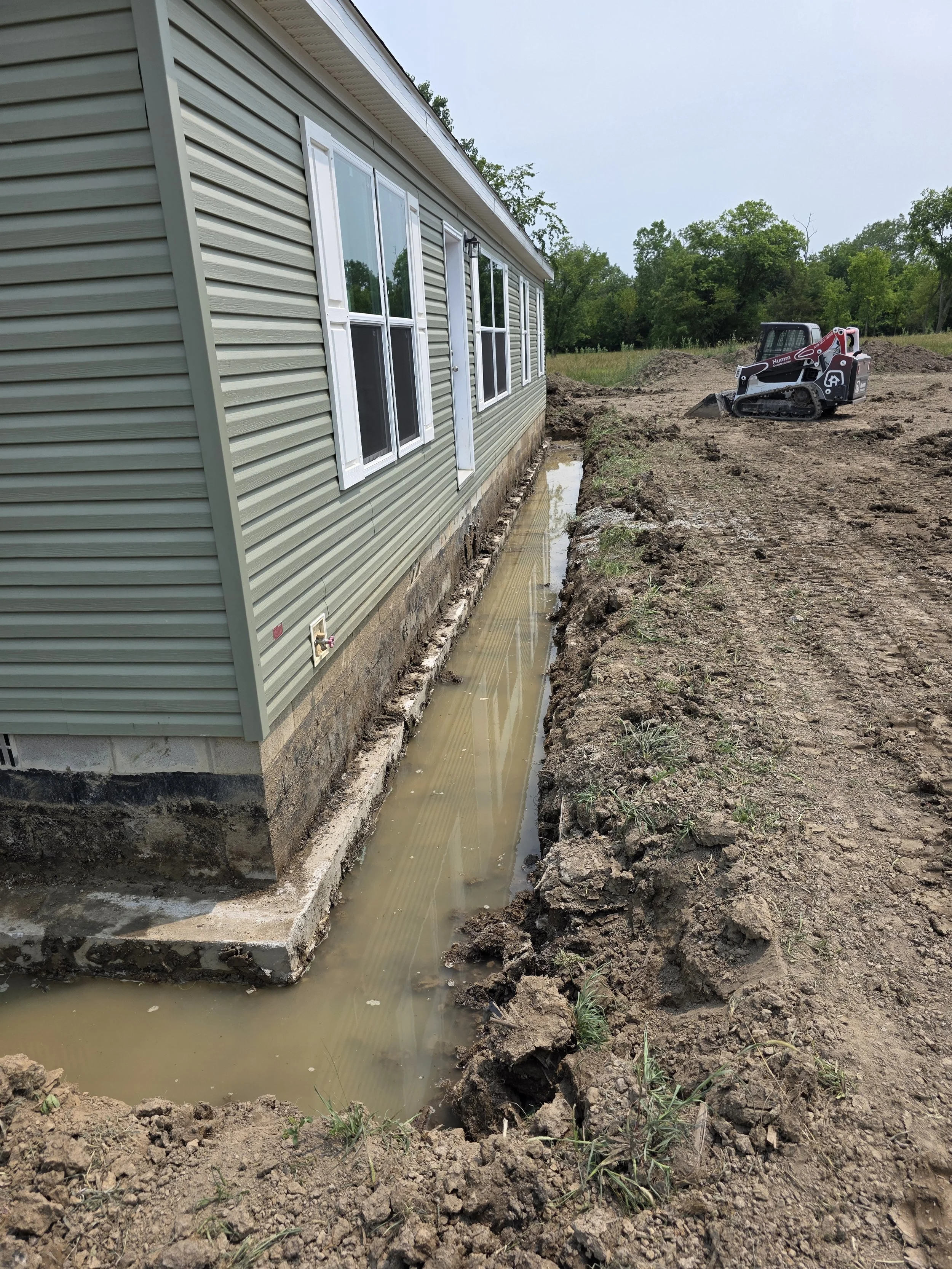 Side view of a house with vinyl siding, and a small excavator on a dirt lot with a trench filled with water along the foundation.