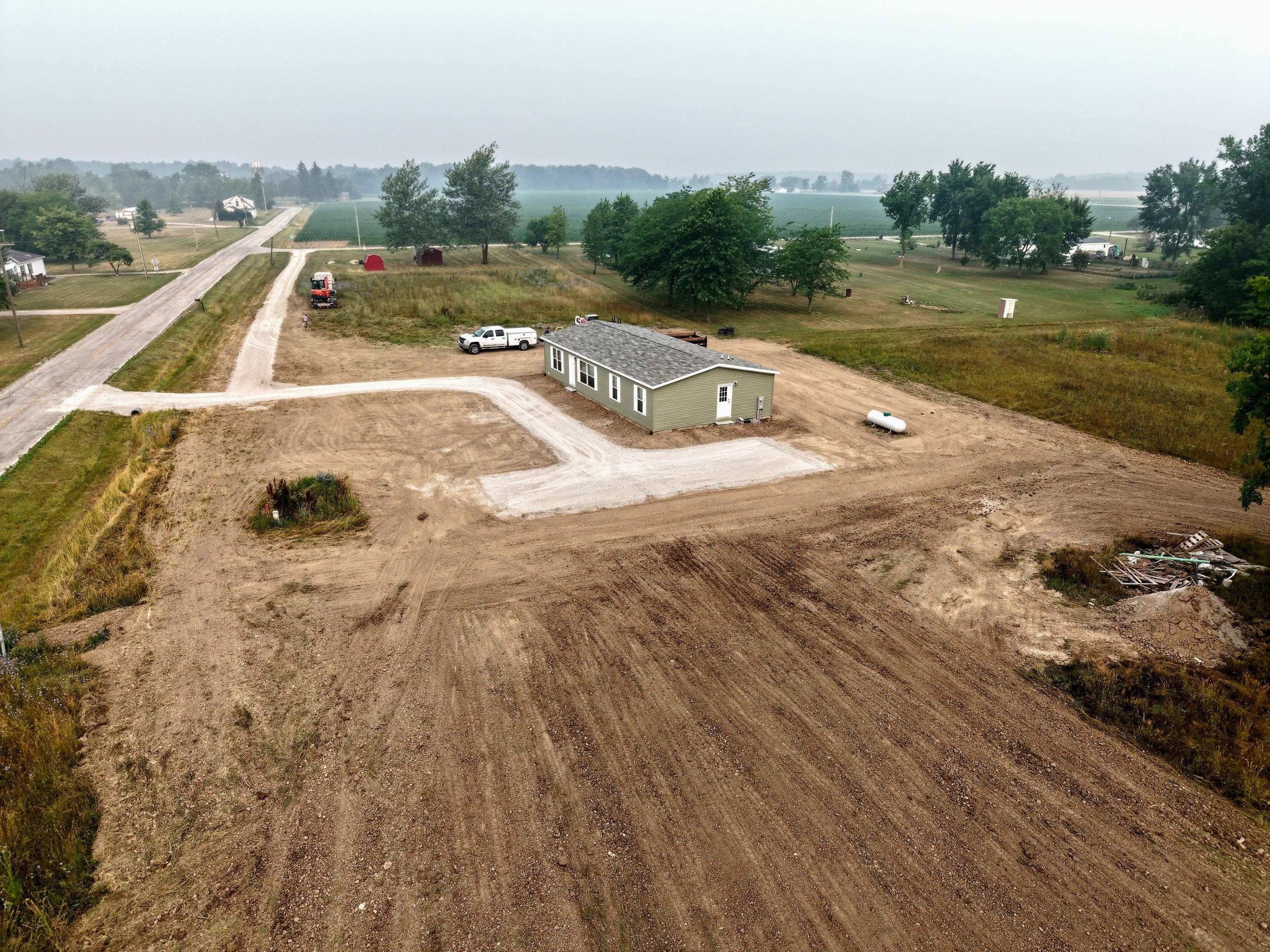 A rural landscape with an under-construction house, a white pickup truck, a red tractor, gravel roads, and lush green trees, with fields and additional houses in the background.
