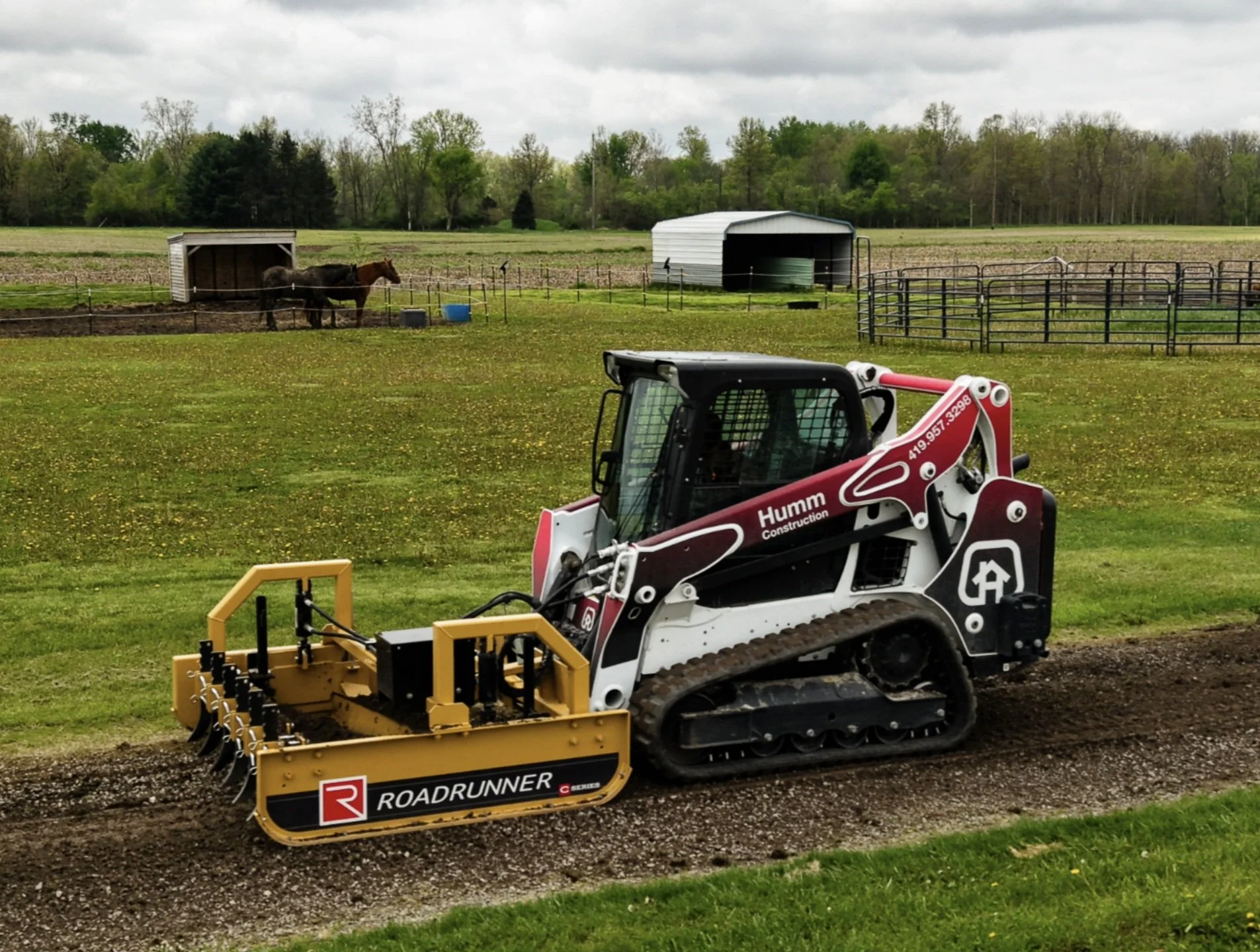 A small tracked tractor with a yellow attachment in a farm setting, with a fenced area containing horses and a horse shelter, alongside open fields and trees under a cloudy sky.