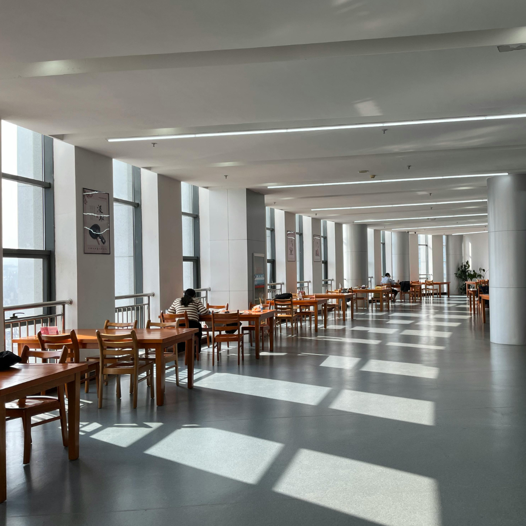 Una sala amplia y luminosa con varias mesas de madera y sillas, con algunas personas sentadas y leyendo. Grandes ventanas dejan entrar luz natural y sombras en el piso.