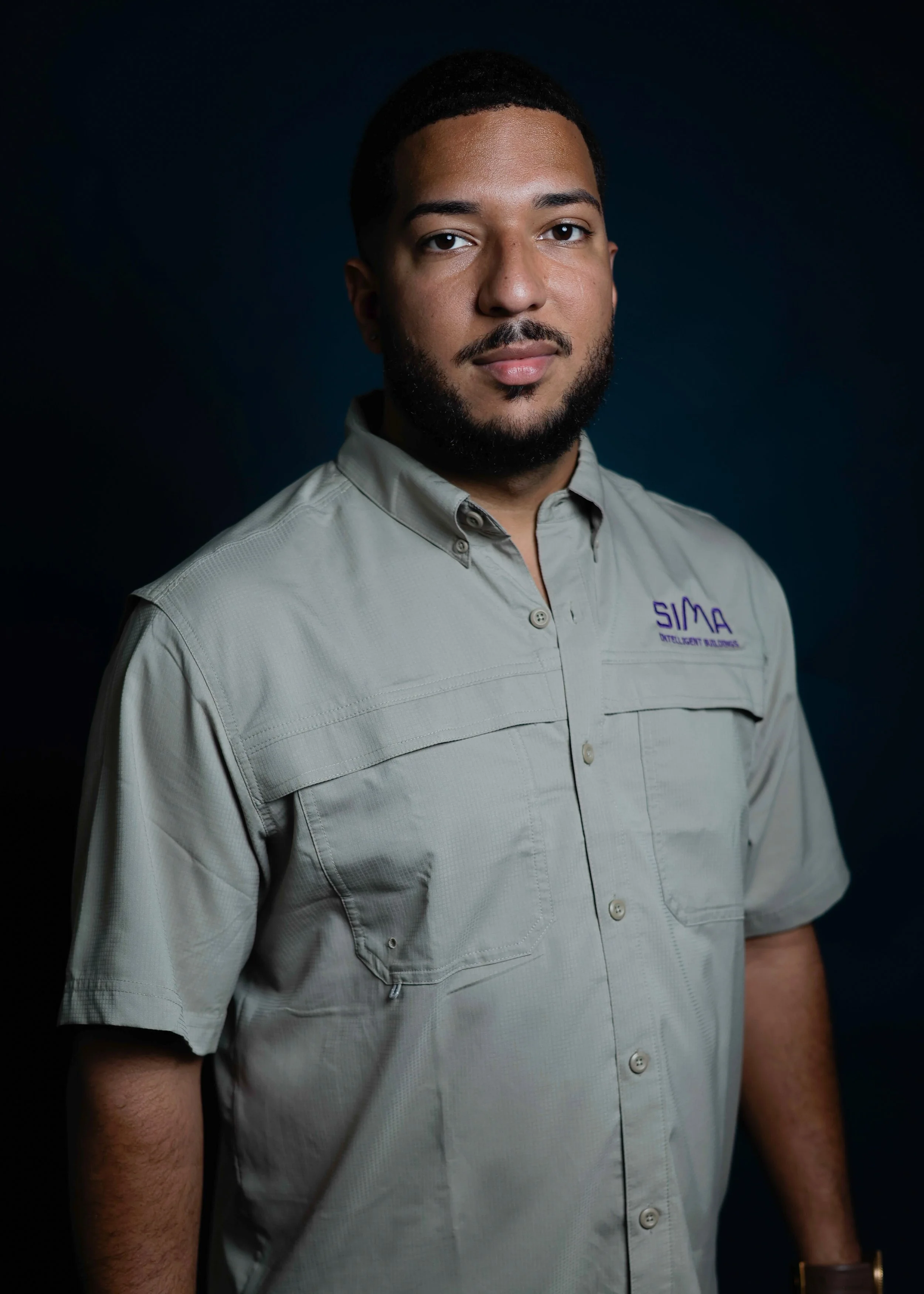 Hombre de piel oscura con barba, cabello corto, con una camisa gris con logotipo 'SIMA' en el pecho, posa de frente contra un fondo oscuro.