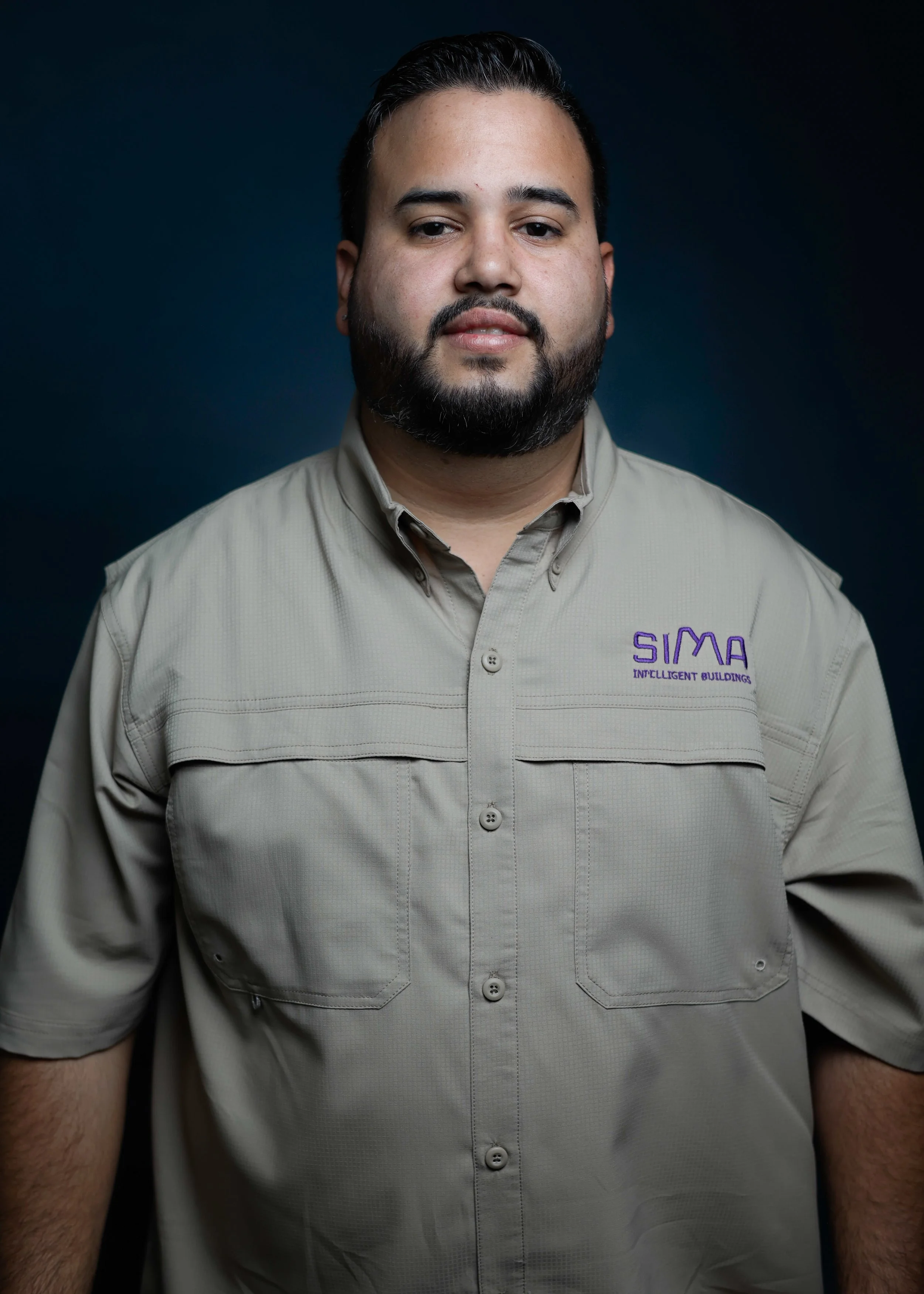 Hombre con barba, cabello oscuro, usa una camisa beige con logotipo en la parte superior izquierda que dice 'SIM A' y 'INLIGERENT BUILDINGS' en color púrpura, en un fondo oscuro.