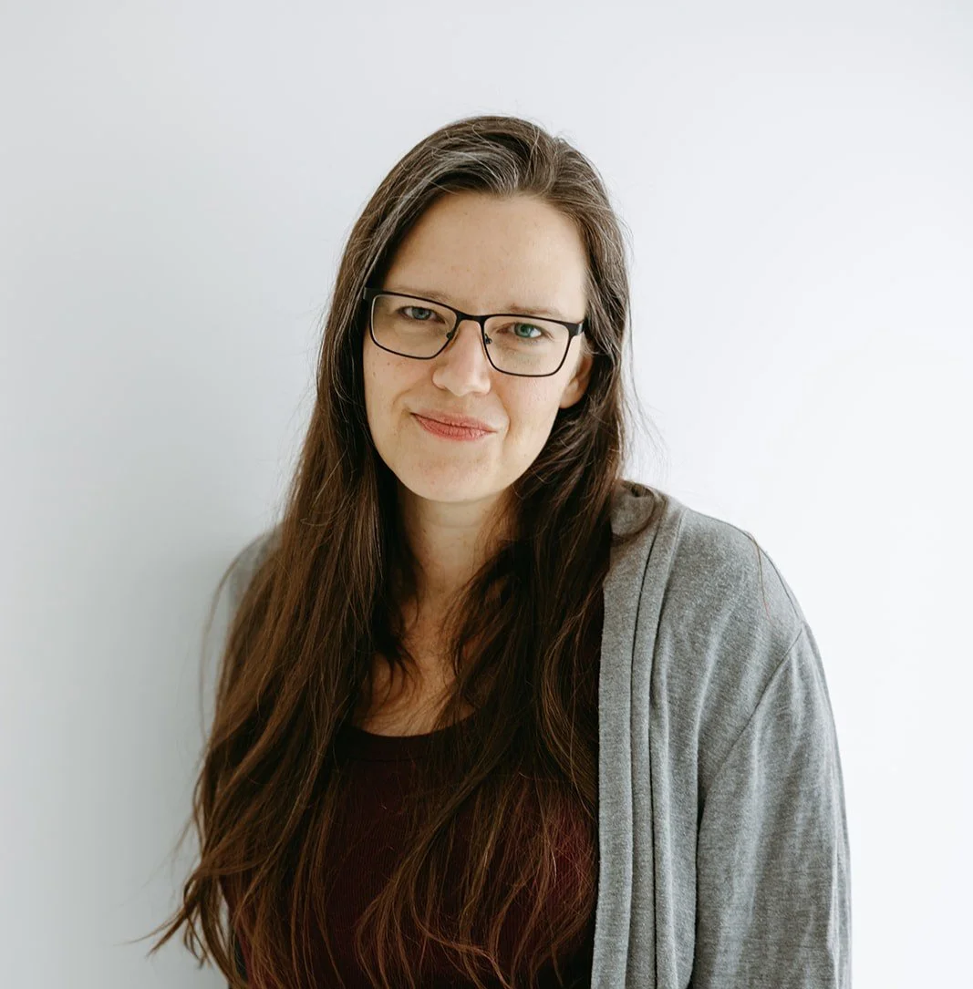 A woman (Nicole Klix) with long brown hair wearing glasses and a gray cardigan, smiling in front of a white background.