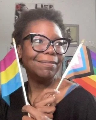 Person holding two small pride flags, one rainbow and one with rainbow and black stripes, in an indoor setting.