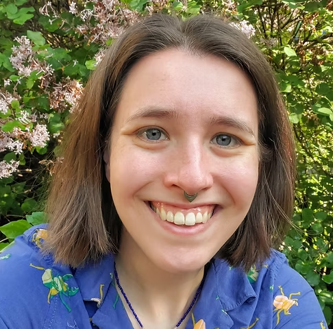 A young woman with shoulder-length brown hair, blue eyes, and a septum piercing smiling outdoors among green foliage and pink flowers.