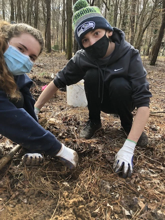Two volunteers smile at the camera while preparing a tree planting site