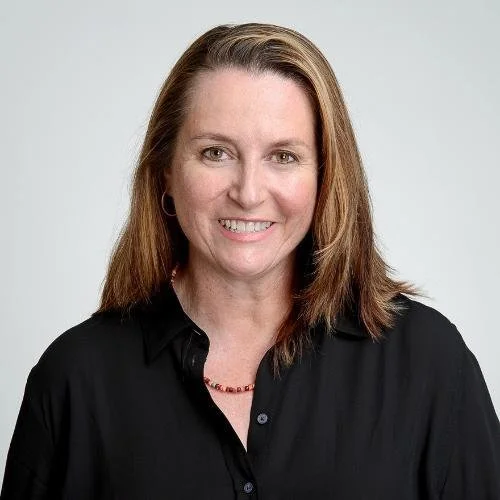 Portrait of a woman with shoulder-length brown hair, wearing a black shirt and a red necklace, smiling at the camera against a light gray background.
