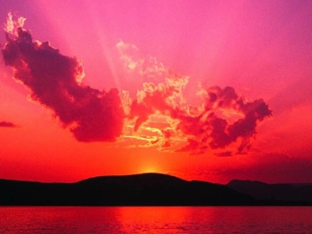 Sunset over a body of water with silhouetted hills in the distance, pink and red sky with dark clouds.