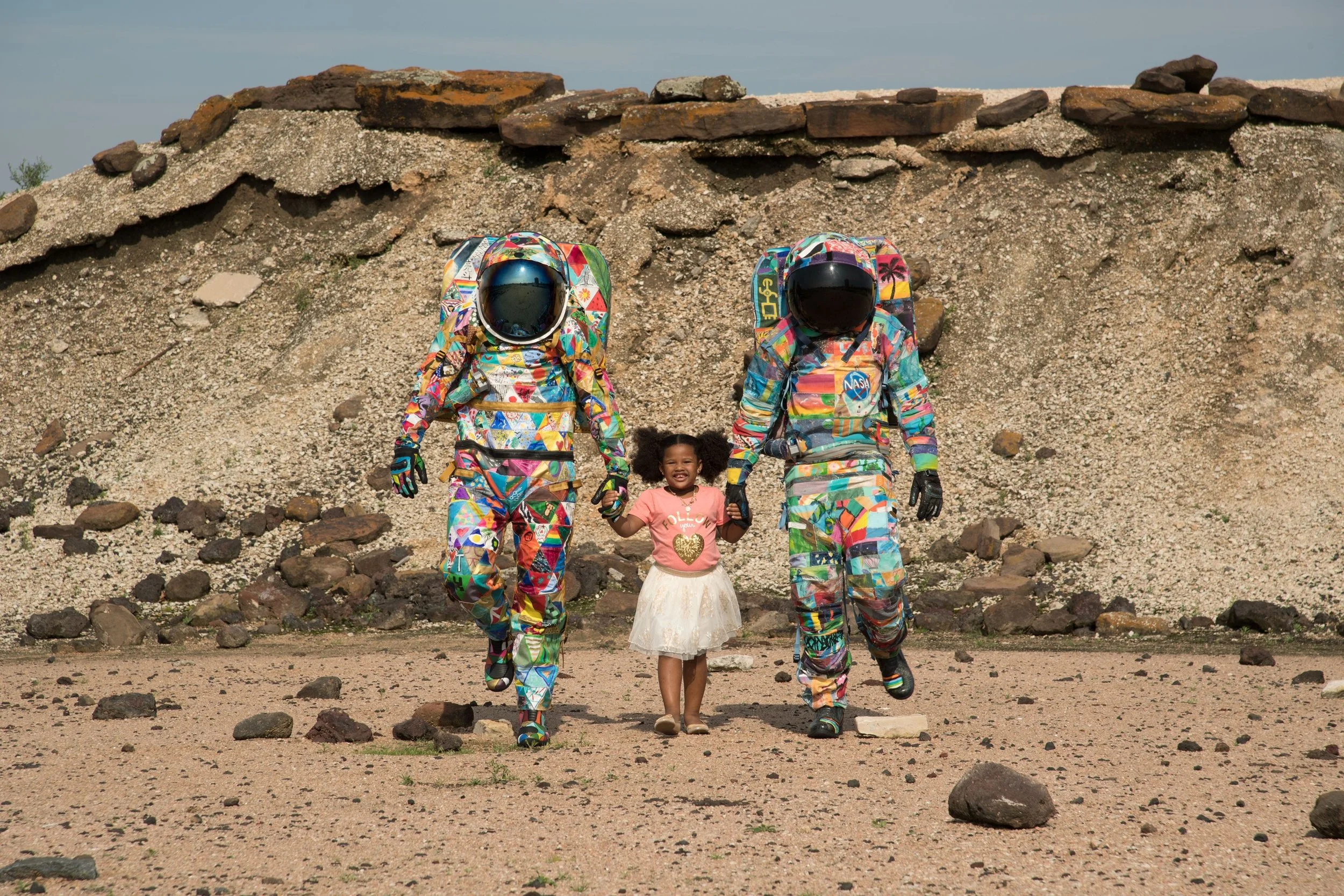 A young girl in a pink shirt and white skirt holding hands with two astronauts in colorful suits with NASA logos, walking on rocky terrain with a dirt mound in the background.