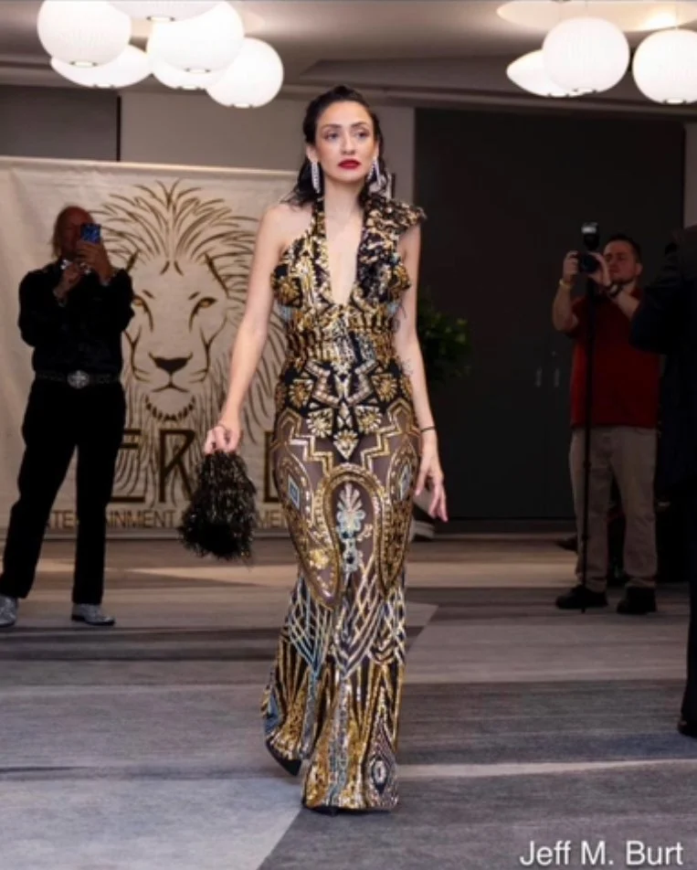 Woman in a patterned, glamorous gown walking on a runway during an indoor event, with photographers taking pictures in the background.