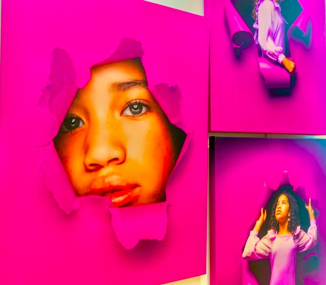 Digital artwork featuring a large, realistic portrait of a young girl's face emerging through a torn pink background, and a smaller girl with curly hair standing in front of a pink background with her hands raised.