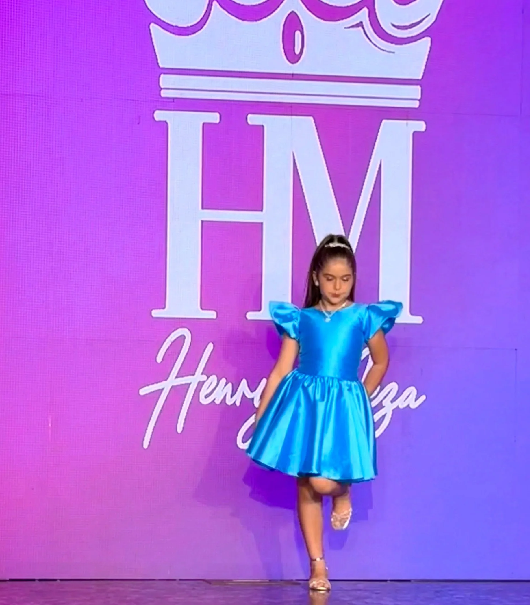 Young girl in a bright blue elegant dress on stage with a pink and purple backdrop that has large white text spelling 'Hermanas' and other cursive writing, performing a dance or pose.