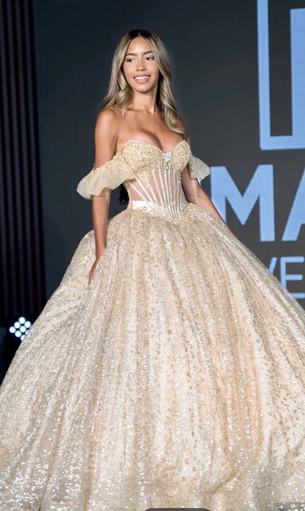 A woman wearing an elegant, off-the-shoulder, beige ball gown with intricate detailing and a full, glittering skirt on a stage with a dark background.