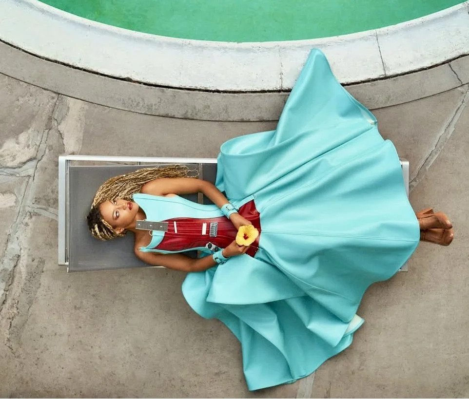 A woman in a bright turquoise ball gown with a low cut red bodice, lying on a metallic lounge chair beside a pool, holding a yellow flower, with blonde braids and makeup.