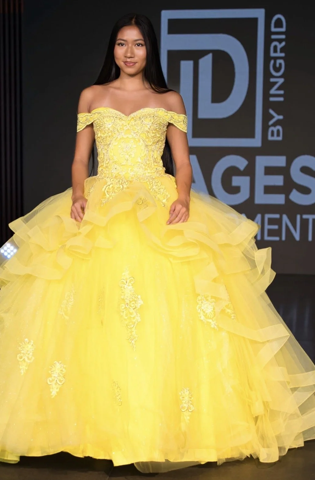 Young woman in a bright yellow ball gown with off-the-shoulder sleeves, intricate embroidery, ruffled layers, and a full skirt, standing in front of a dark background with logo and text.