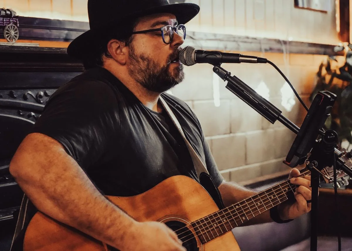 A man with glasses, a black hat, and a beard is playing an acoustic guitar and singing into a microphone, with a tablet mounted on a stand in front of him.