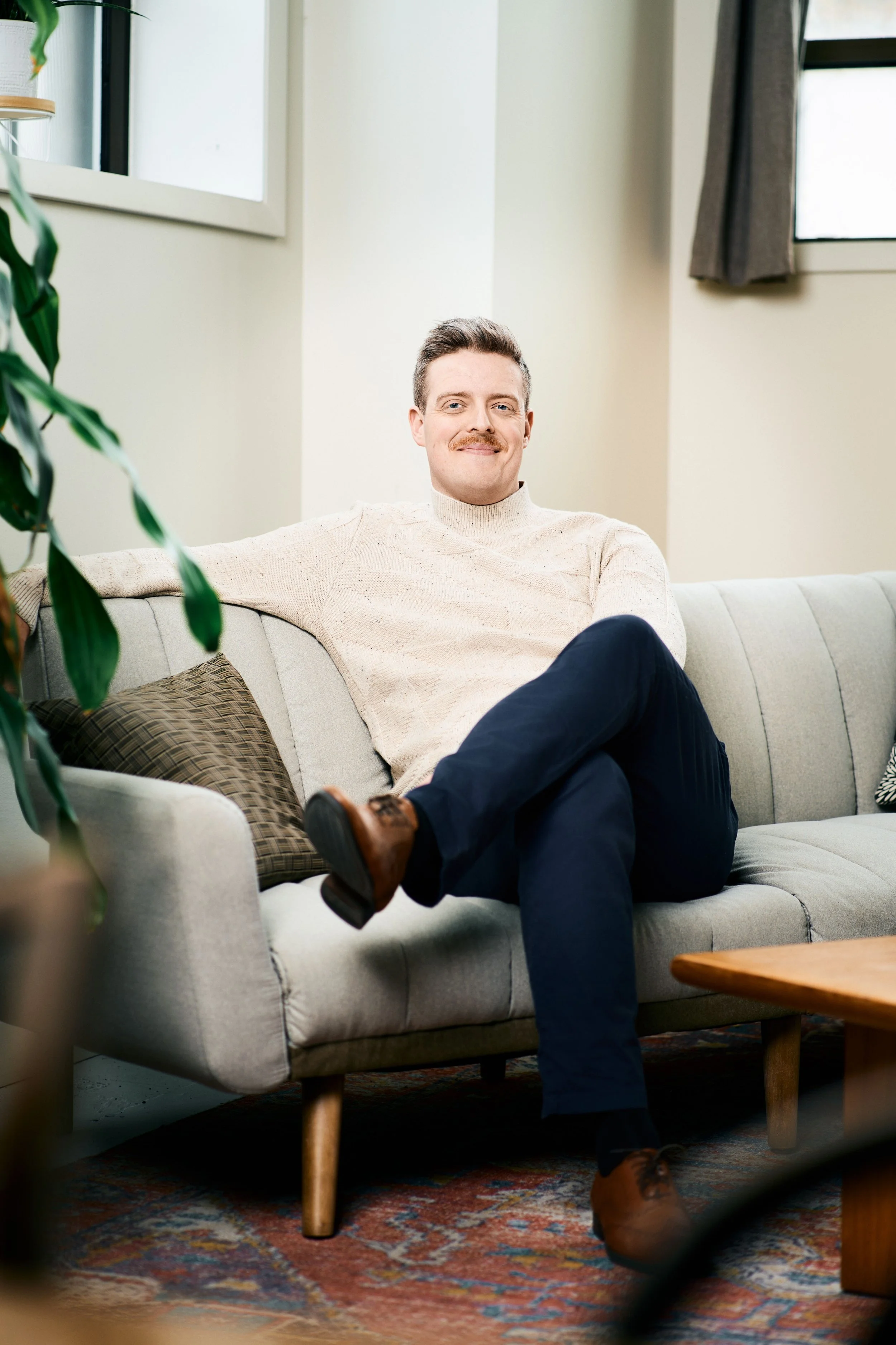A man with a mustache sitting on a beige couch in a living room, smiling, with his legs crossed and wearing a light sweater, dark pants, and brown boots. There is a window with curtains behind him, and a patterned rug on the floor.
