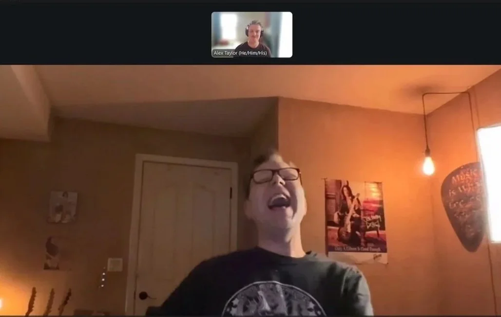 A young person with glasses singing or shouting with eyes closed in a room with posters and a hanging light bulb.