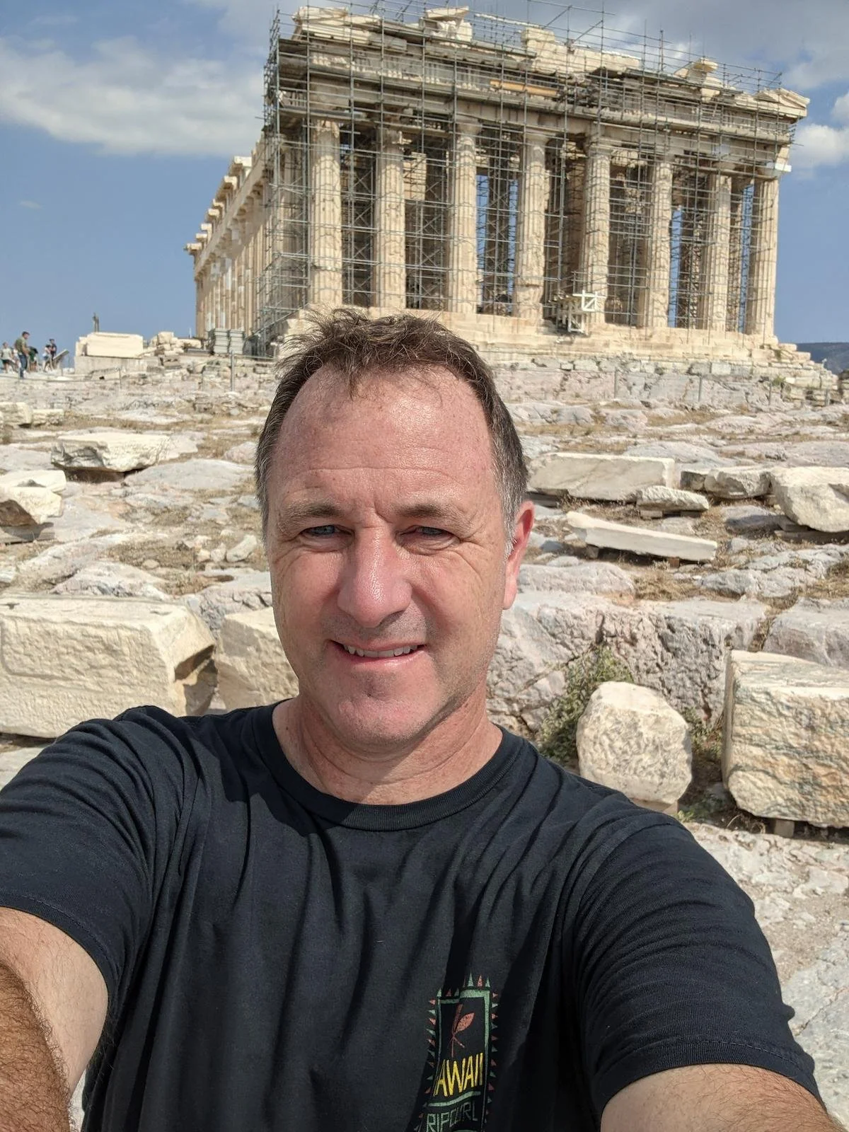 A man taking a selfie with the Parthenon in Athens, Greece, in the background. The Parthenon is under restoration scaffolding, and there are scattered rocks and ruins on the ground.